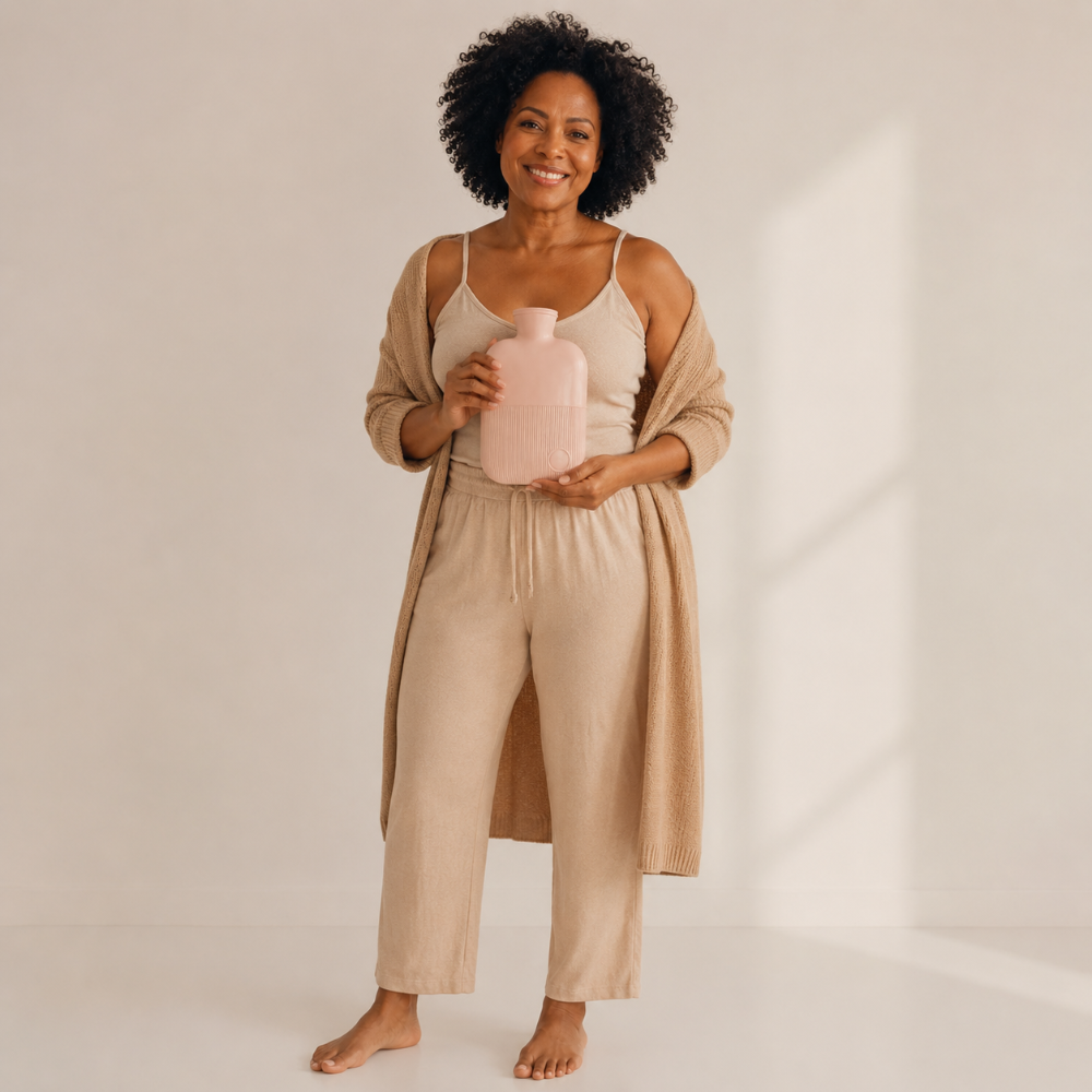 A woman stands barefoot in a bright studio setting, wearing a beige camisole, matching drawstring pants, and a draped cardigan, while holding a pale pink bottle.